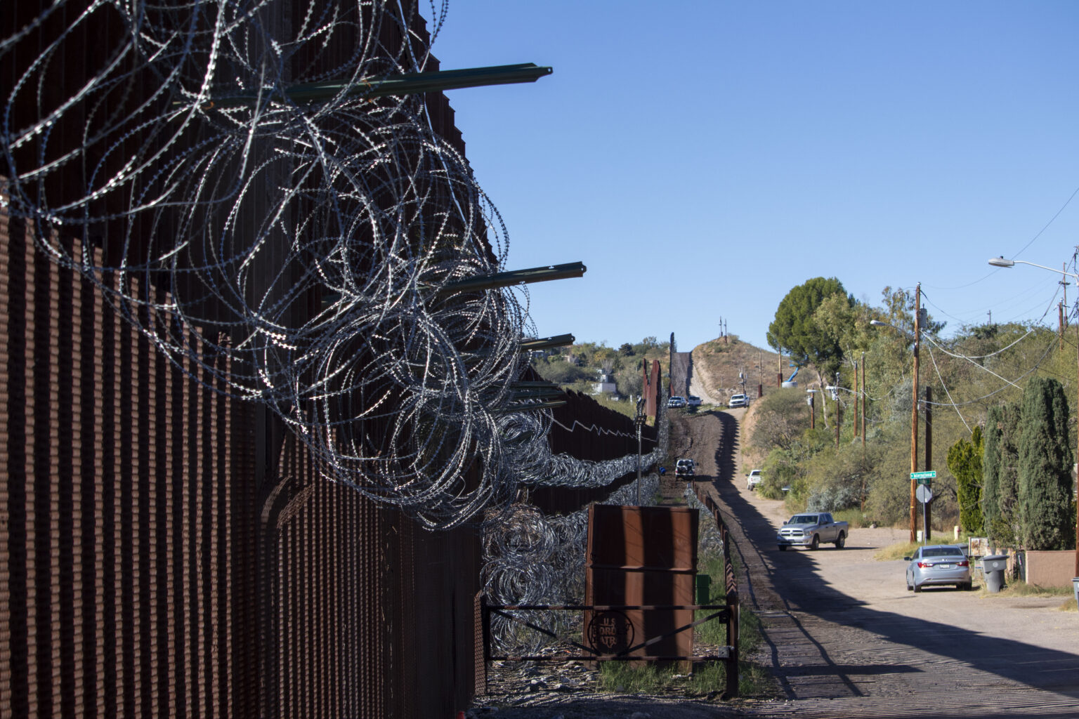 Razor wire tops a section of the U.S.-Mexico border fence with a nearby road in view — a visual representation of the border-security context for the new trooper funding.
