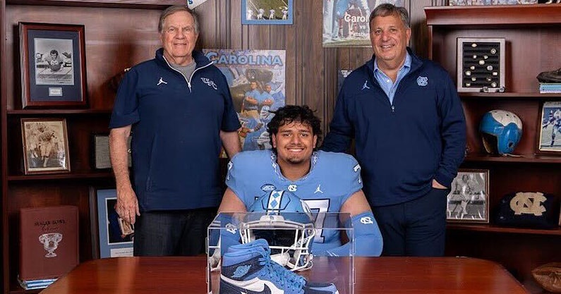Signing-style photo of the committed lineman in North Carolina gear, seated with Tar Heels helmet and sneakers on display after announcing his commitment to UNC.