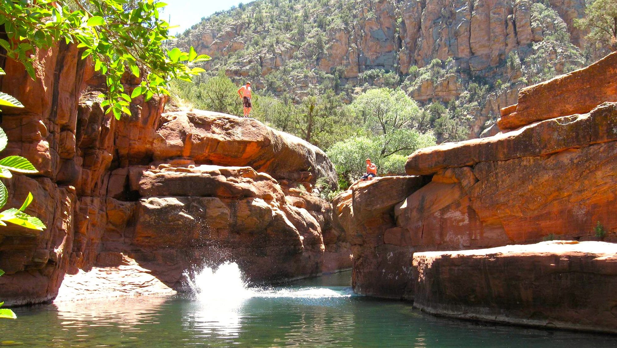 People jump from sandstone ledges into a clear canyon pool — a popular swim spot in Arizona’s red‑rock country.