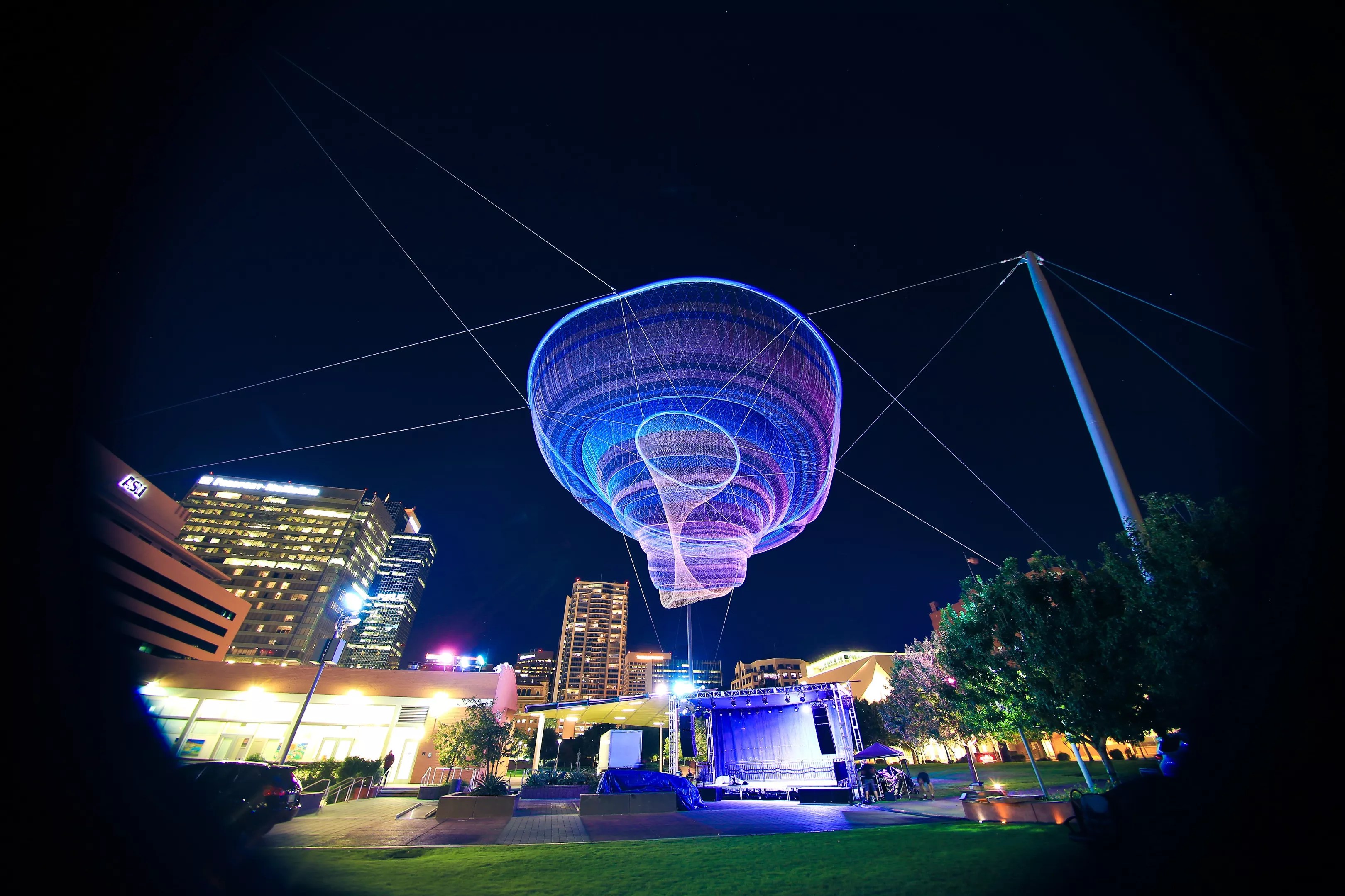 Illuminated suspended public‑art installation over a downtown outdoor stage, shown lit at night and listed as a free outdoor attraction this weekend.