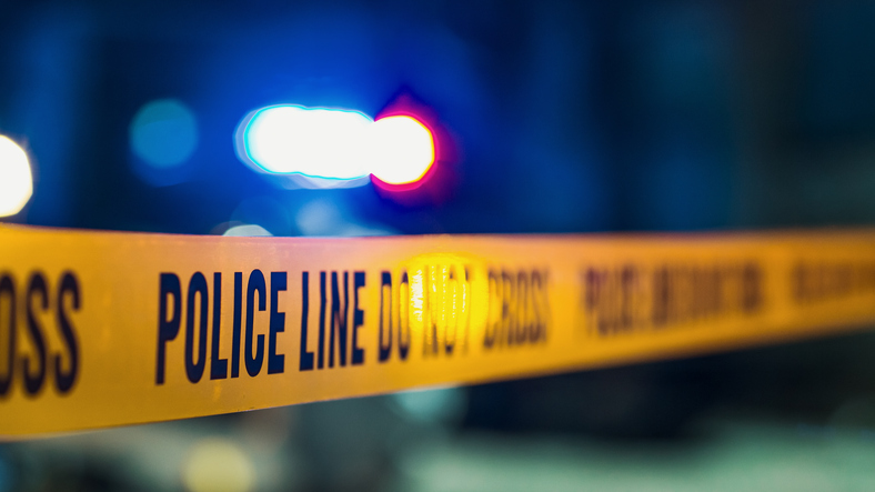 Yellow 'POLICE LINE DO NOT CROSS' tape with flashing patrol lights in the background — scene imagery tied to the Arizona State Troopers' investigation of the May 25 shooting near Benson, Arizona.