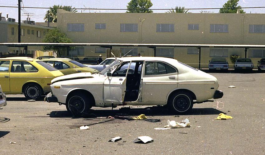 Color photo of the damaged white sedan and scattered debris at the parking‑lot scene of the 1976 car‑bombing referenced in the story.