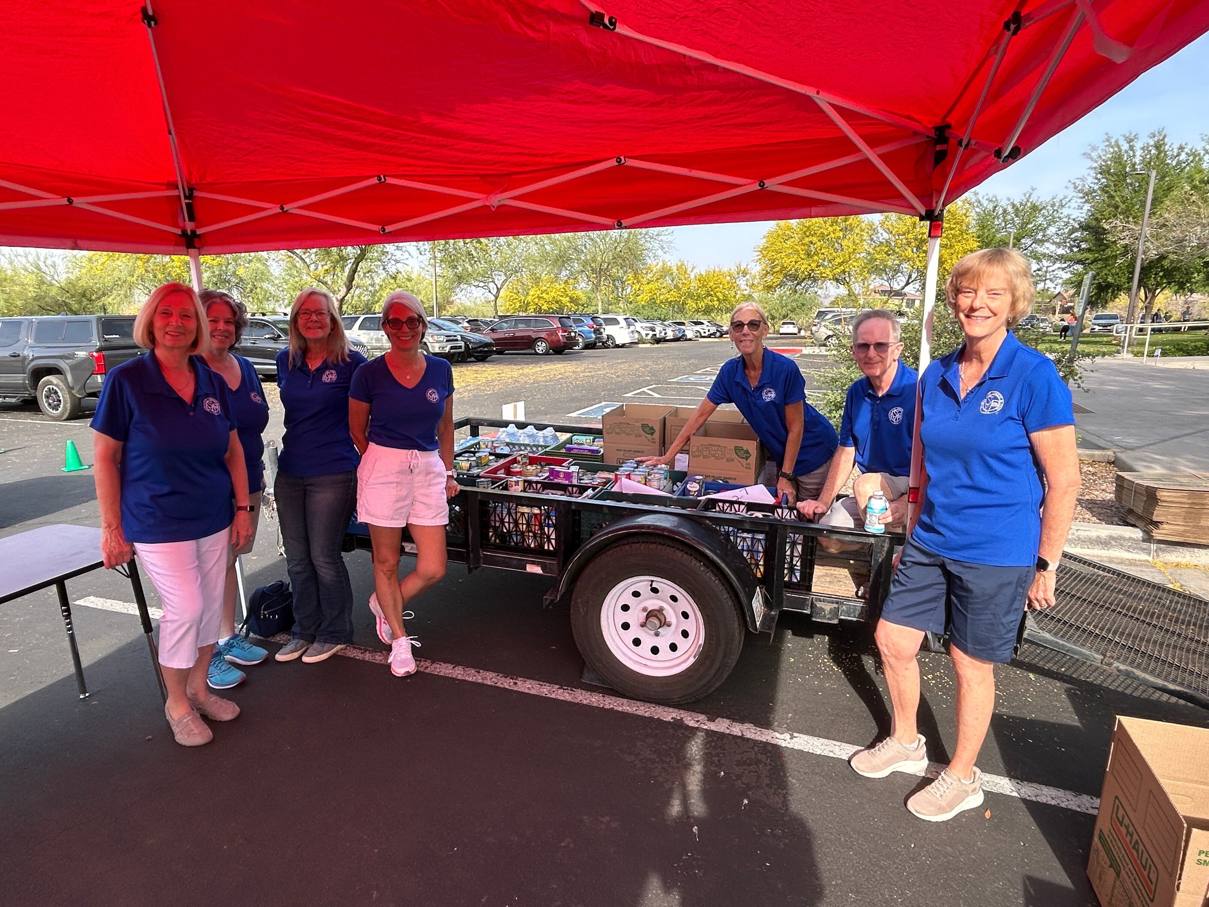 Volunteers in matching shirts stand behind a trailer loaded with canned goods and boxed donations at the event collection point.