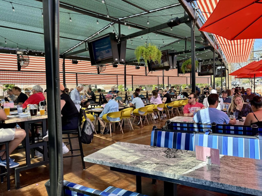 A busy, shaded outdoor patio with multiple TVs and patrons watching a match — representative of the lively bars and restaurants recommended for cheering on the U.S. in the World Cup.
