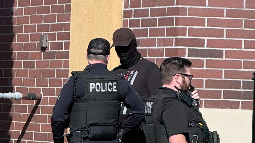 Police officers speaking with a masked individual outside a brick building — an illustrative image of an arrest or custody encounter linked to reporting on a trooper's alleged use of force and subsequent probe.